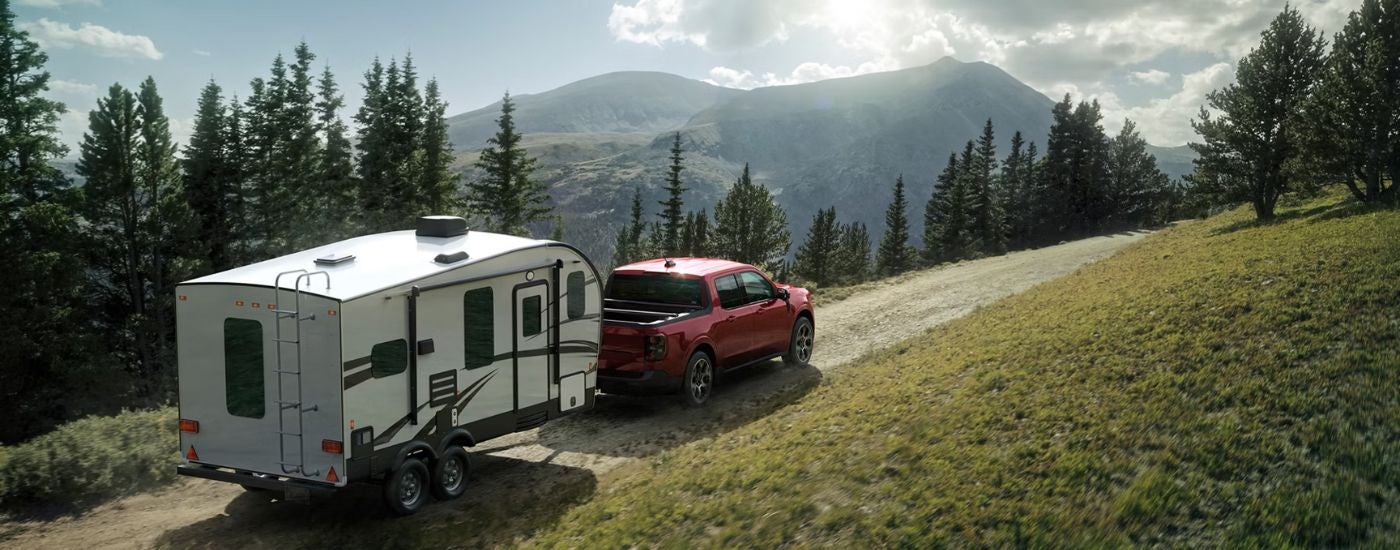 A red 2026 Ford Maverick shown towing a camper down a trail.